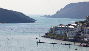 SALCOMBE 2 This landscape photograph captures the coastal town of Salcombe in Devon, England, during a bright afternoon in early spring. Salcombe’s waterfront buildings are prominently featured along the water’s edge, with the main subject being the town itself, nestled in a picturesque setting on the southern coast of the United Kingdom. The image shows the calm waters of the estuary, marked with navigation poles, and is framed by the rugged, tree-covered hills that slope down to the coast, characteristic of Devon’s dramatic shoreline. The clear lighting suggests mild spring weather and highlights the architectural details of the waterfront properties and hotels that define Salcombe’s unique charm. The photograph also reveals the confluence of natural beauty and local life, as boats and construction activity on one of the buildings hint at ongoing development in this popular English seaside destination.